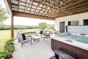 A covered outdoor patio with a hot tub, seating area, and a view of the countryside.