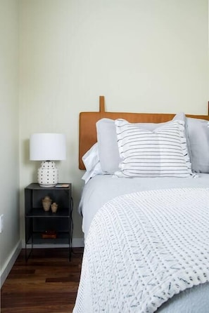 A neatly made bed with a wooden headboard, a white knitted throw, and a bedside table with a lamp and decorative items.