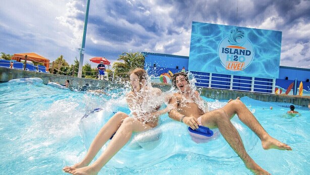 A swimming pool with people enjoying the water, a large sign reading "ISLAND 2 LIVE!", and a building with blue and white colors in the background. - Next Door To It All! Disney/Dining & More mins away 2Bed 2Bth Condo (Kissimmee)