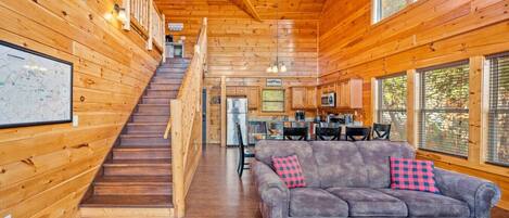 A wooden cabin interior with a staircase leading to a loft bedroom, a living area with a sofa and a coffee table, and a kitchen area.