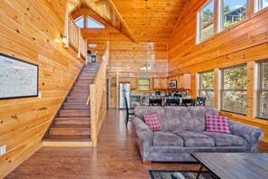 A wooden cabin interior with a staircase leading to a loft bedroom, a living area with a sofa and a coffee table, and a kitchen area.