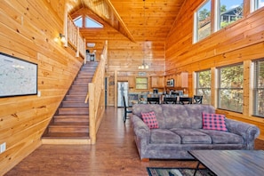 L’intérieur d’une cabane en bois comprend un escalier menant à une chambre à coucher située en mezzanine, un espace de vie avec un canapé et une table basse, ainsi qu’une cuisine.