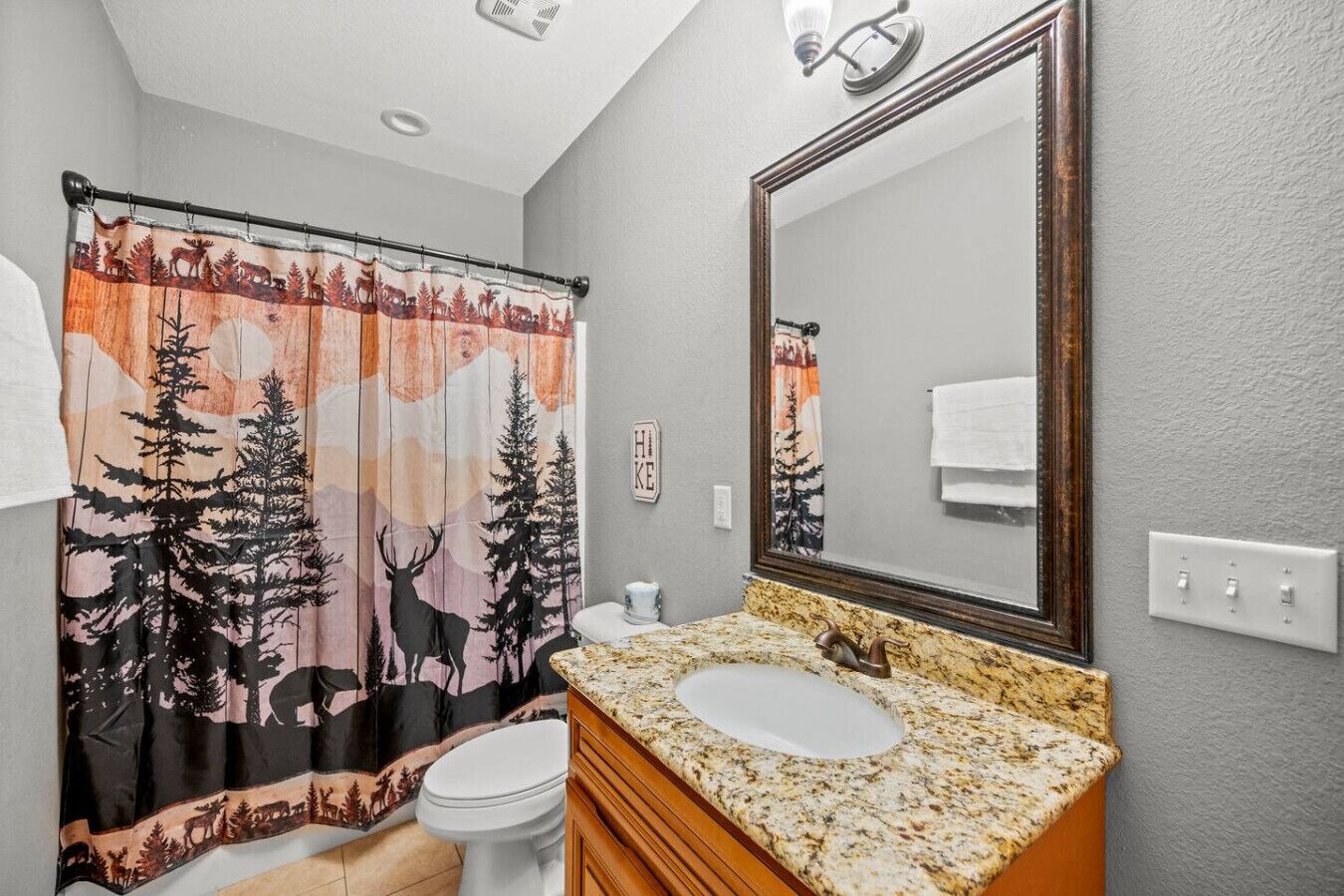 A bathroom with a shower curtain featuring a forest and deer design, a white toilet, a granite sink, and a large mirror.