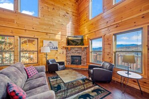 A cozy cabin interior with a fireplace, wooden walls, and large windows offering scenic views. - Luxe Pool Cabin w/MTN views! Hot Tub + Theater (Pigeon Forge)
