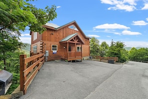 A log cabin with a porch, a fenced driveway, and a view of the surrounding forest.