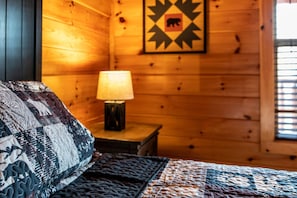A wooden cabin room with a bed, a nightstand with a lamp, and a wall with a bear symbol.