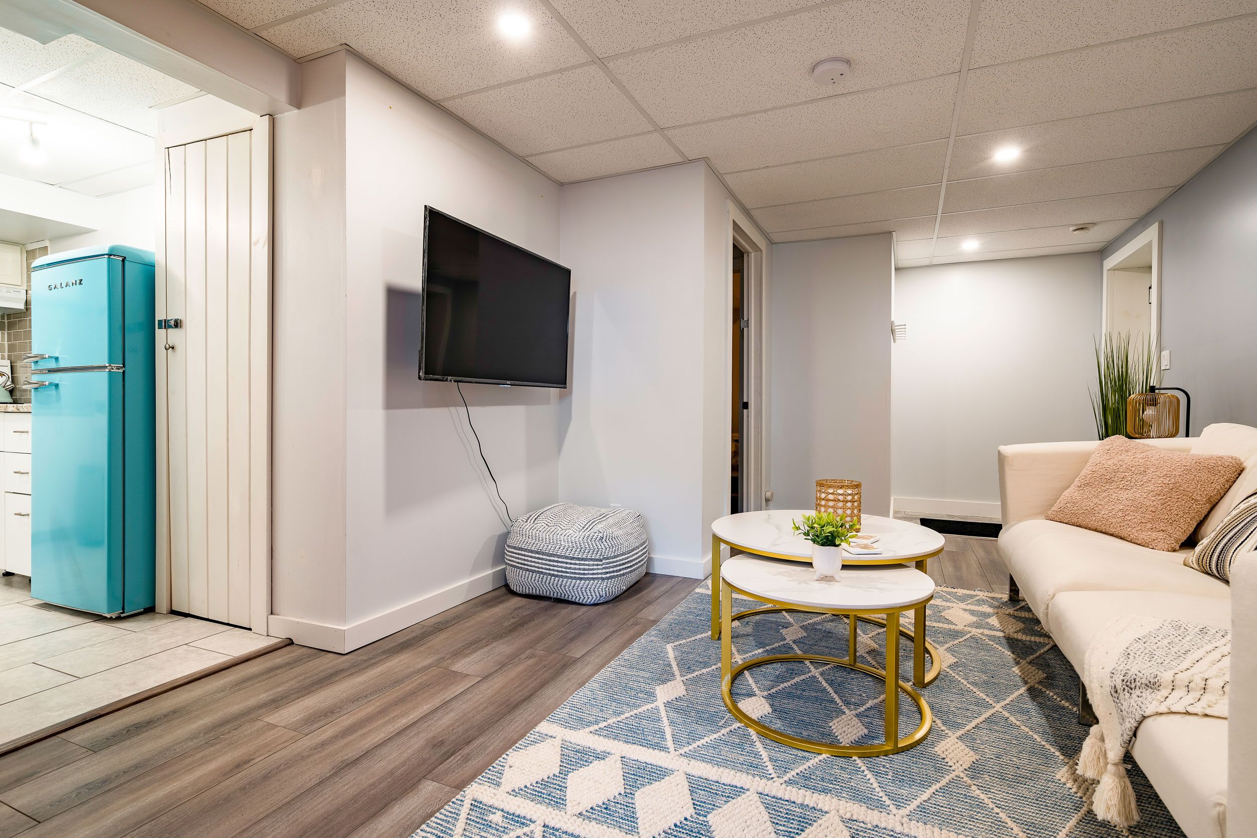 A modern living room with a teal refrigerator, a flat-screen TV mounted on the wall, a round coffee table with gold legs, and a beige sofa with a patterned throw.