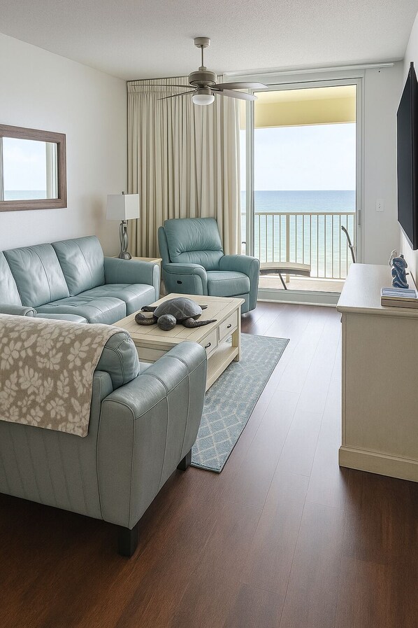 A modern living room with a sofa, armchairs, a coffee table, and a TV. There is a balcony with a view of the ocean.
