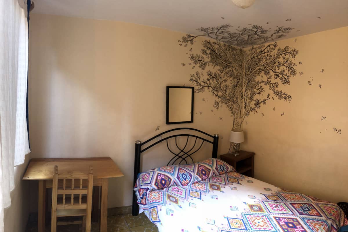 Makuuhuoneessa on sänky, puinen tuoli, pieni pöytä, yöpöytä ja seinämaalaus.