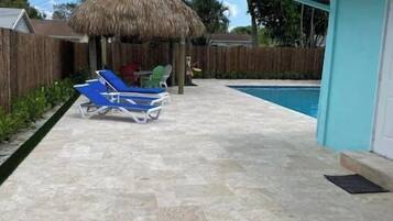 Un patio trasero con piscina, una estructura de techo de paja y un sillón de descanso azul.
