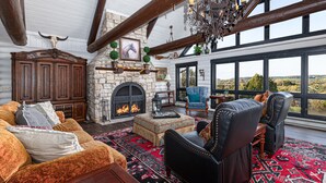 A cozy living room with a fireplace, leather chairs, a patterned rug, and a large window overlooking a scenic landscape. - WOW! Unique Galena Dream Cabin. Hot Tub, Games, Fire Pit, Fully Stocked. (Galena)