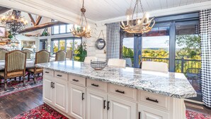 A spacious kitchen with granite countertops, white cabinetry, and a large island. There are two chandeliers with antler chandeliers, a dining area with a table and chairs, and a view of the outdoors through large windows. - WOW! Unique Galena Dream Cabin. Hot Tub, Games, Fire Pit, Fully Stocked. (Galena)