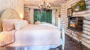 A cozy bedroom with a large bed, a fireplace, and a scenic view through the window. - WOW! Unique Galena Dream Cabin. Hot Tub, Games, Fire Pit, Fully Stocked. (Galena)