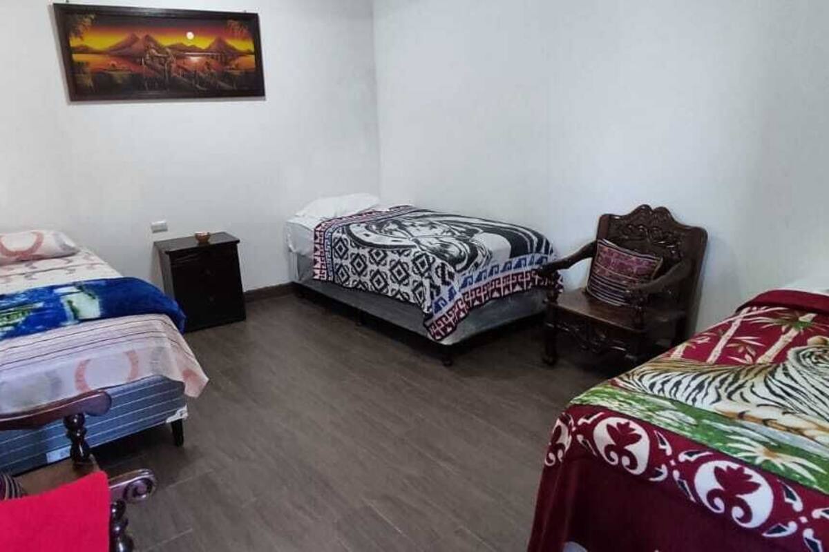 Una habitación pequeña y funcional con dos camas individuales, una silla de madera, una mesita y un estante en la pared.