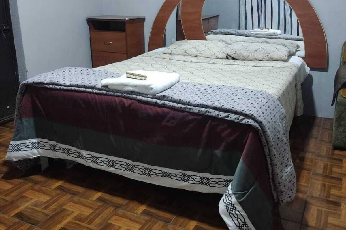 Un dormitorio con suelo de madera, una cama con colcha estampada, una mesita de noche de madera y un aparador.