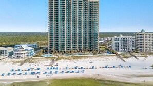 A tall hotel with a red roof, surrounded by a beach with blue umbrellas and a pool. - Phoenix West 2207 Upscale Luxury | Updated | Beachfront | Waterslide-Lazy River (Orange Beach)