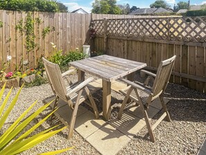 Un conjunto de mesa y sillas de madera sobre un patio de grava, con una cerca de madera y plantas al fondo.