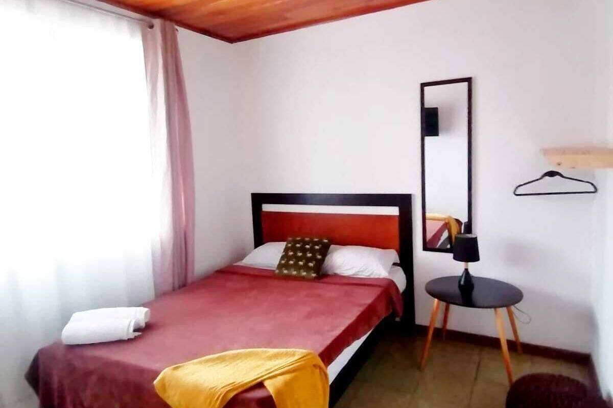 A bedroom with a wooden ceiling, a bed with a red bedspread, a bedside table with a lamp, and a mirror on the wall.