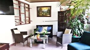Un salon avec un canapé, des fauteuils, une table basse et un téléviseur. On y trouve également des plantes et un tableau accroché au mur.