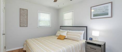 Un dormitorio con cama, mesita de noche, ventilador de techo y dos ventanas.