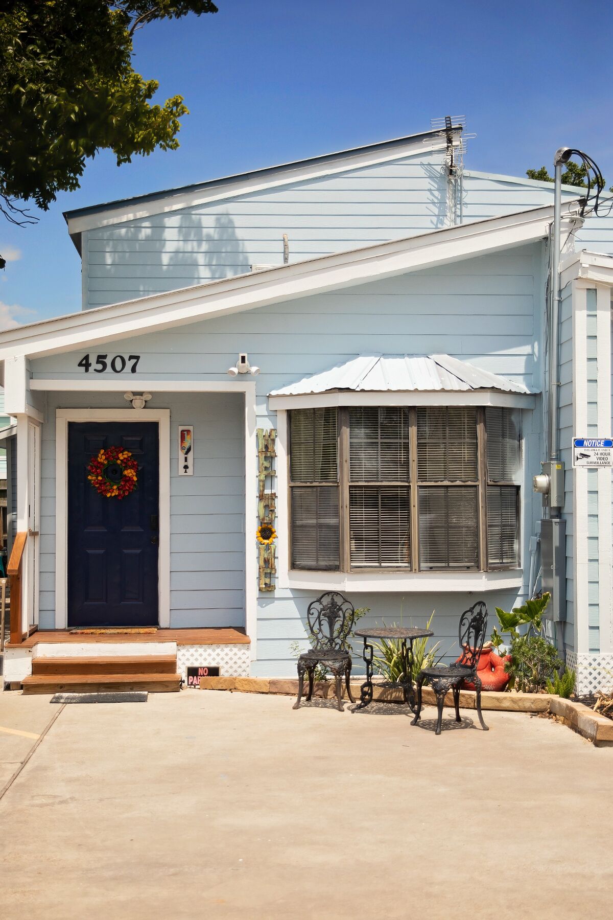 A light blue house with a dark blue door, a number '4507' above the door, a small porch with steps, and a table with two chairs outside.
