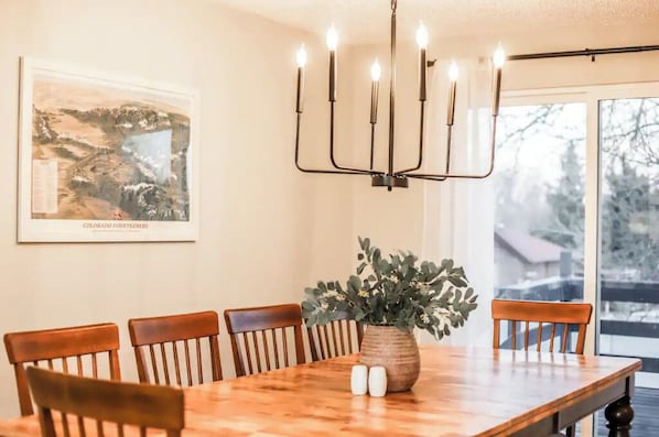 A dining room with a wooden table, six chairs, a candelabra with lit candles, a framed landscape painting, and a potted plant.