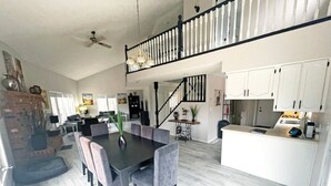 A spacious living area with a dining table, grey upholstered chairs, a staircase, and a kitchen area with white cabinets.