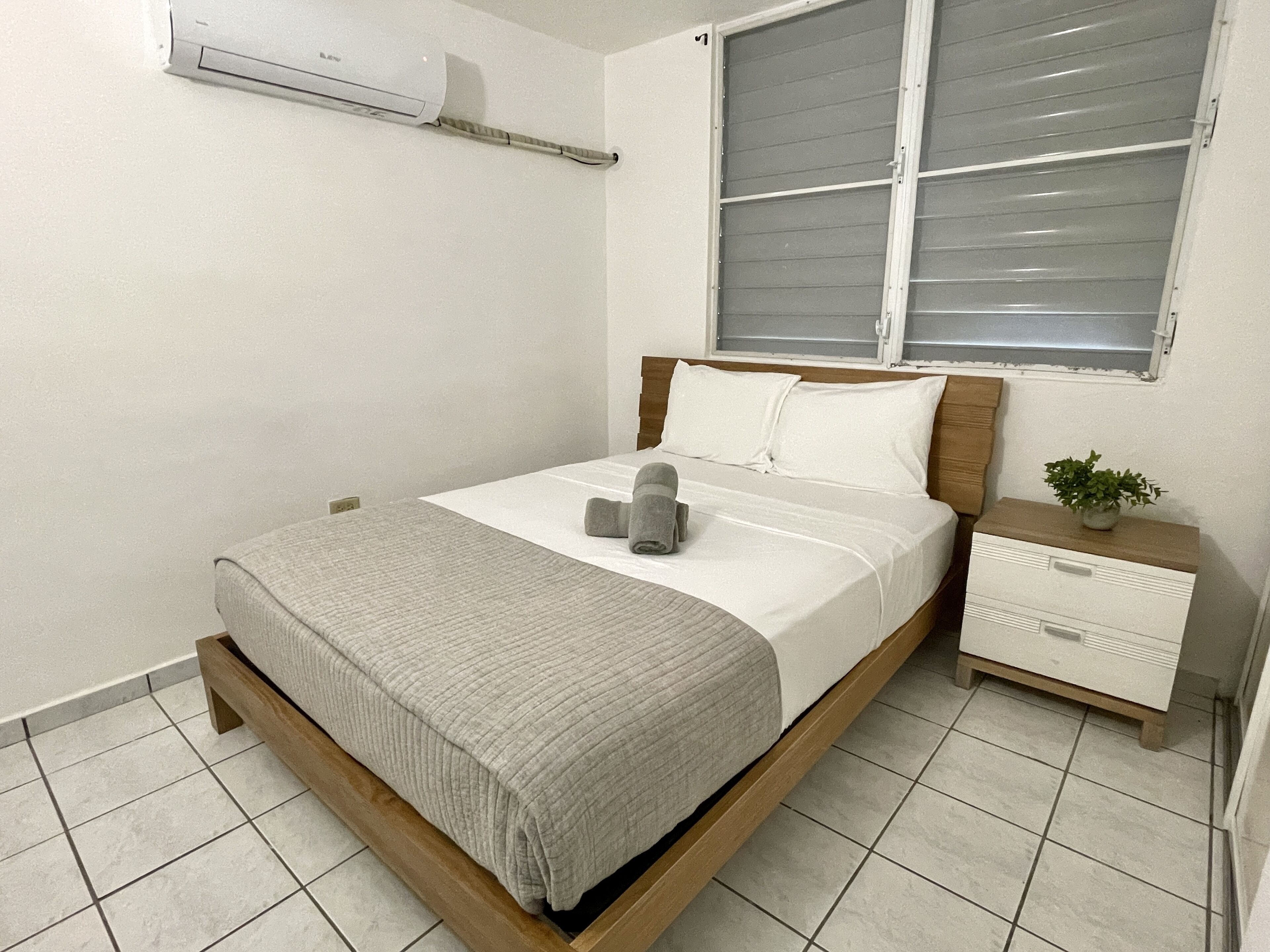 Una cama bien tendida con un edredón gris, una mesita de noche con una planta y una ventana con persianas.