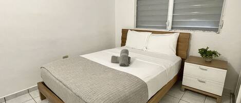 Una cama bien tendida con un edredón gris, una mesita de noche con una planta y una ventana con persianas.