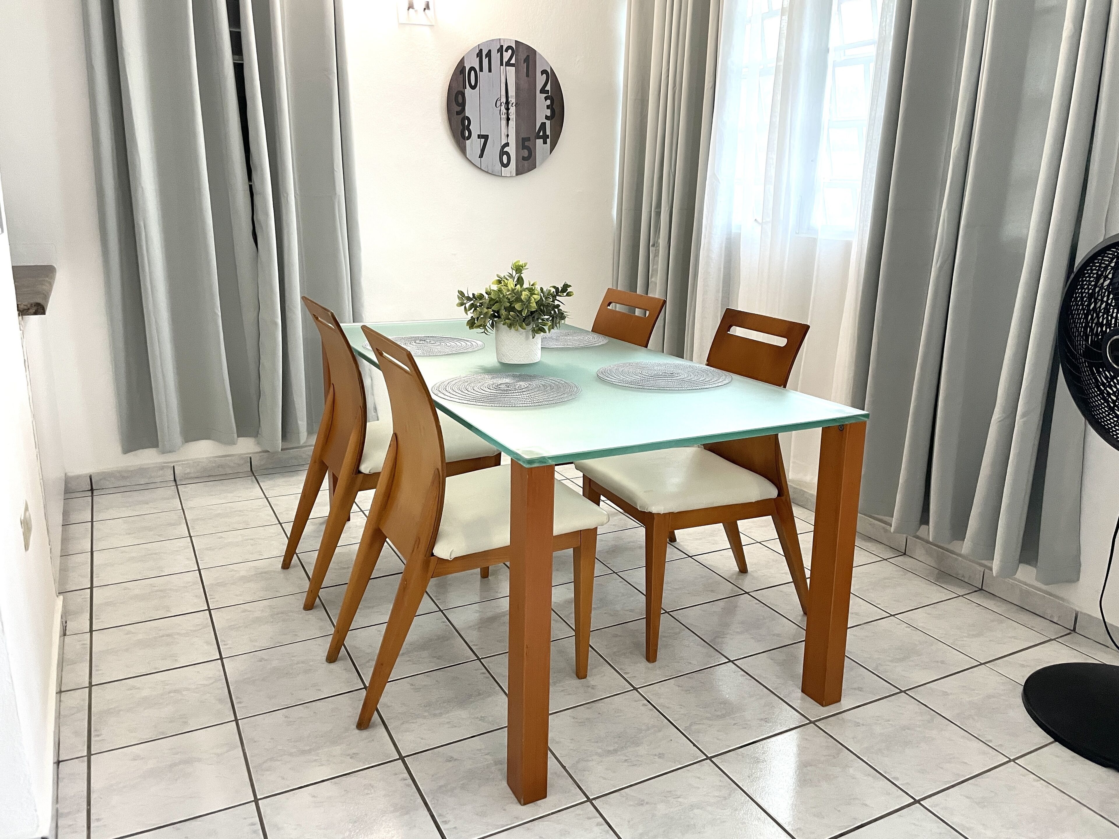 A dining area with a glass-top table, wooden chairs, a potted plant, and a wall clock.