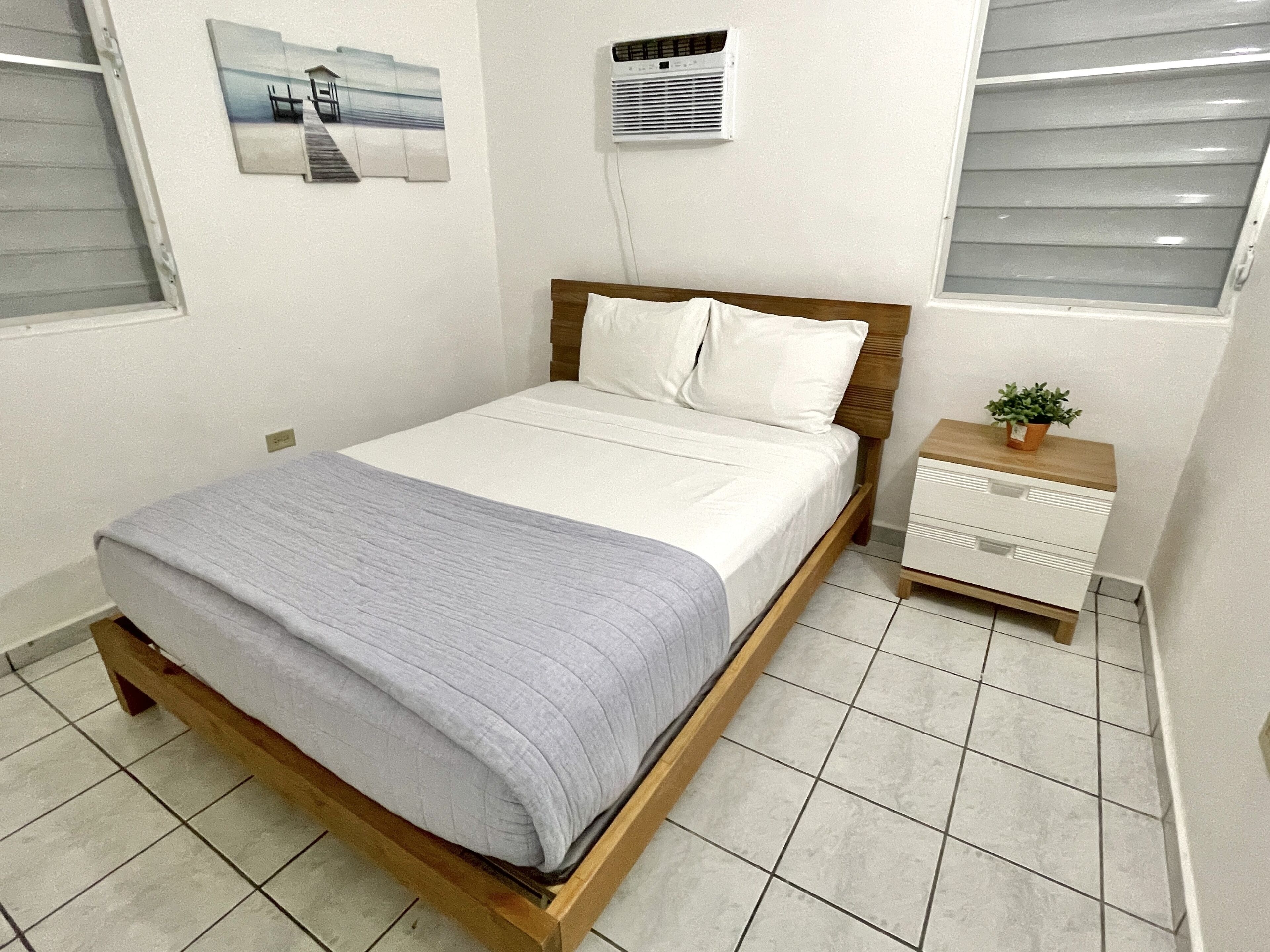 Una cama bien hecha con estructura de madera y una manta gris, una mesita de noche con una planta, y un aire acondicionado instalado en la pared.