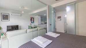 Ein modernes Hotelzimmer mit einem großen Bett, einer Sitzecke, einem Fernseher und einer Glaskonstruktion, die den Wohnbereich von der Küche trennt.