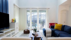 A living room with a sofa, coffee table, and TV. There is a balcony with a glass door and a Coca-Cola refrigerator.