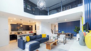 A spacious living area with a blue sofa, a wooden coffee table, and a dining table with chairs. A kitchen is visible in the background, and there is a balcony with a railing.