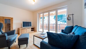 Ein modernes Wohnzimmer mit Sofa, Sessel, Hocker und einem Fernseher auf einer Holzkommode.