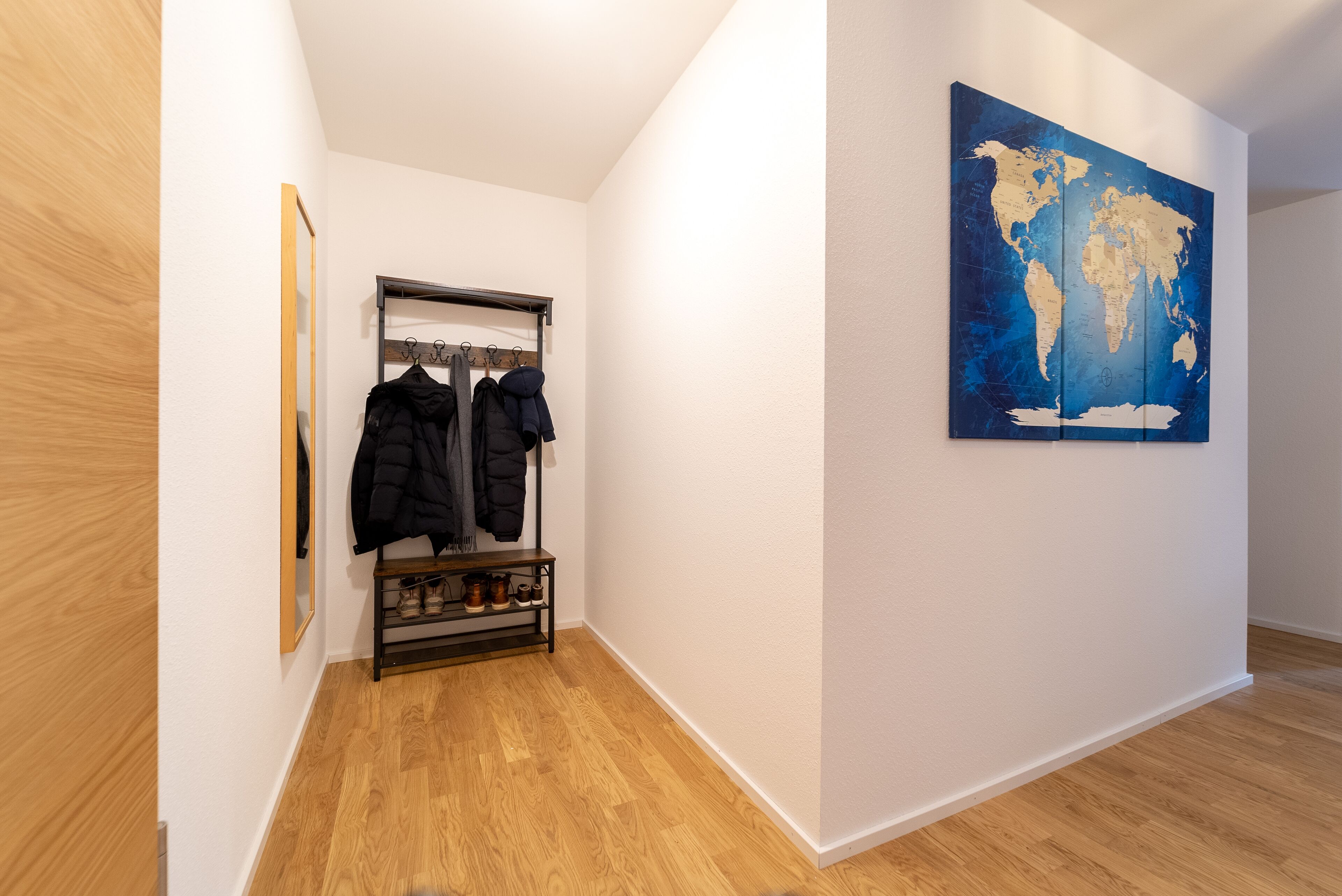 Un couloir avec un porte-manteau sur lequel sont accrochées plusieurs vestes, un meuble à chaussures rempli de chaussures et une grande carte du monde accrochée au mur.