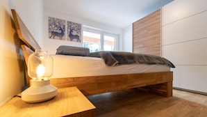 Ein Schlafzimmer mit einem hölzernen Bett, einem Nachttisch mit Lampe und einem Fenster mit Blick auf Bäume.