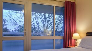 Camera d'albergo con un letto, un comodino con una lampada e una finestra che si affaccia su alberi e una recinzione.