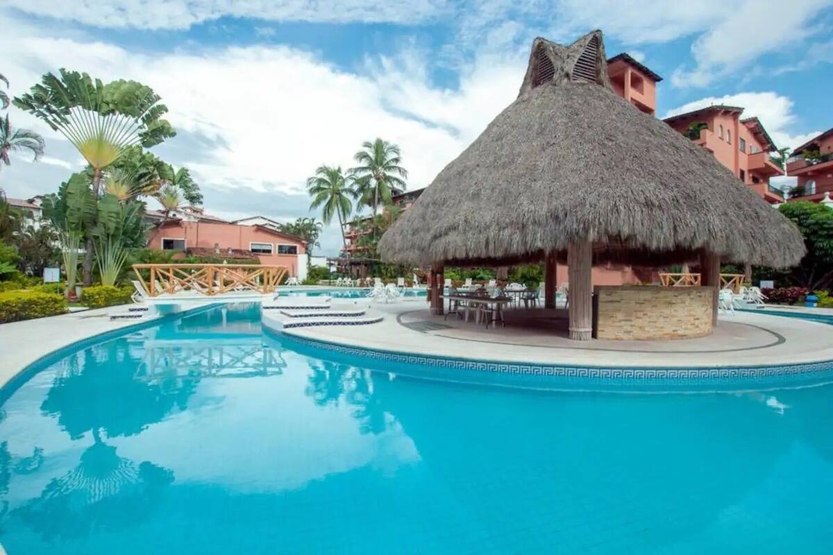 A resort pool with a thatched-roof structure, palm trees, and pink buildings in the background.