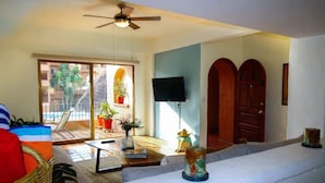 A living room with a ceiling fan, a TV mounted on the wall, a balcony with potted plants, and a large window. - CONDO PSOL BY TRVL2HM (Puerto Vallarta)