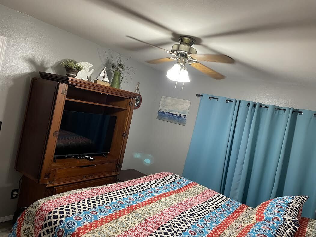 Une chambre à coucher avec un lit, une armoire en bois, un ventilateur de plafond et une fenêtre avec des rideaux bleus.