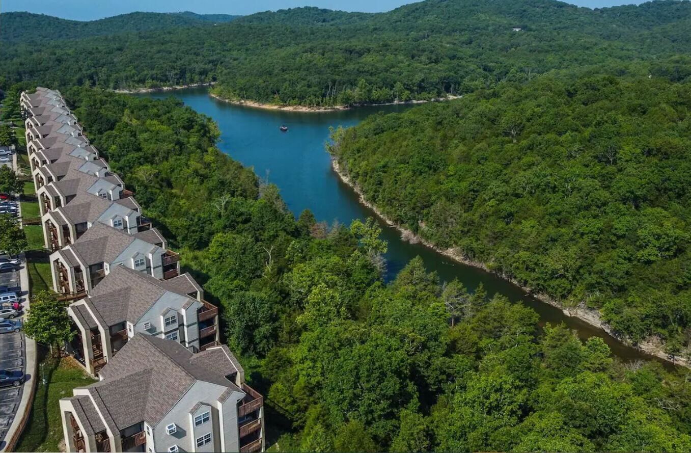 Zona residencial con una hilera de casas a lo largo de un río, rodeada de un bosque denso.