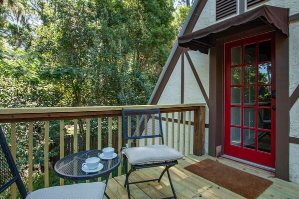 A wooden deck with two chairs and a table, a red door, and a white building with brown trim.