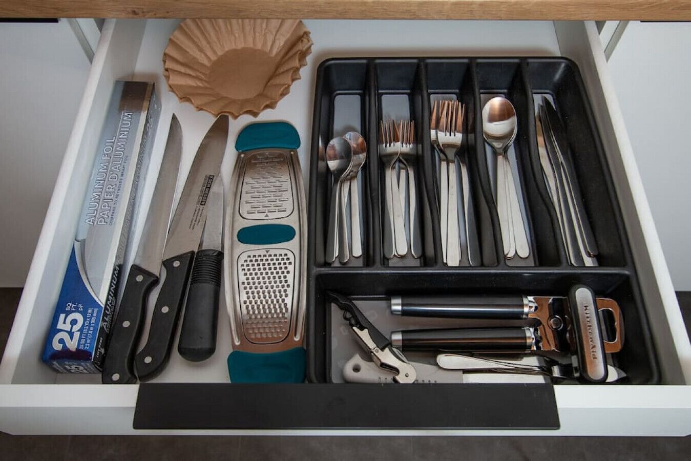 Un cajón con varios utensilios de cocina, como un rallador, cucharas, tenedores y un cuchillo.