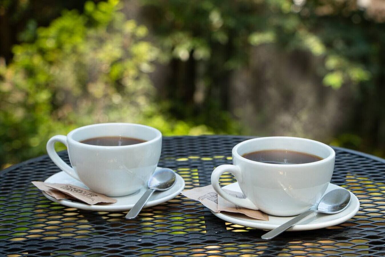 Deux tasses de café blanc sont posées sur une table ronde en métal, accompagnées d’une cuillère. La scène se déroule en plein air, avec une végétation luxuriante en arrière-plan.