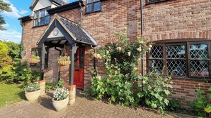 Une maison en briques avec une porte rouge, un petit porche et un jardin où poussent différentes plantes et fleurs.