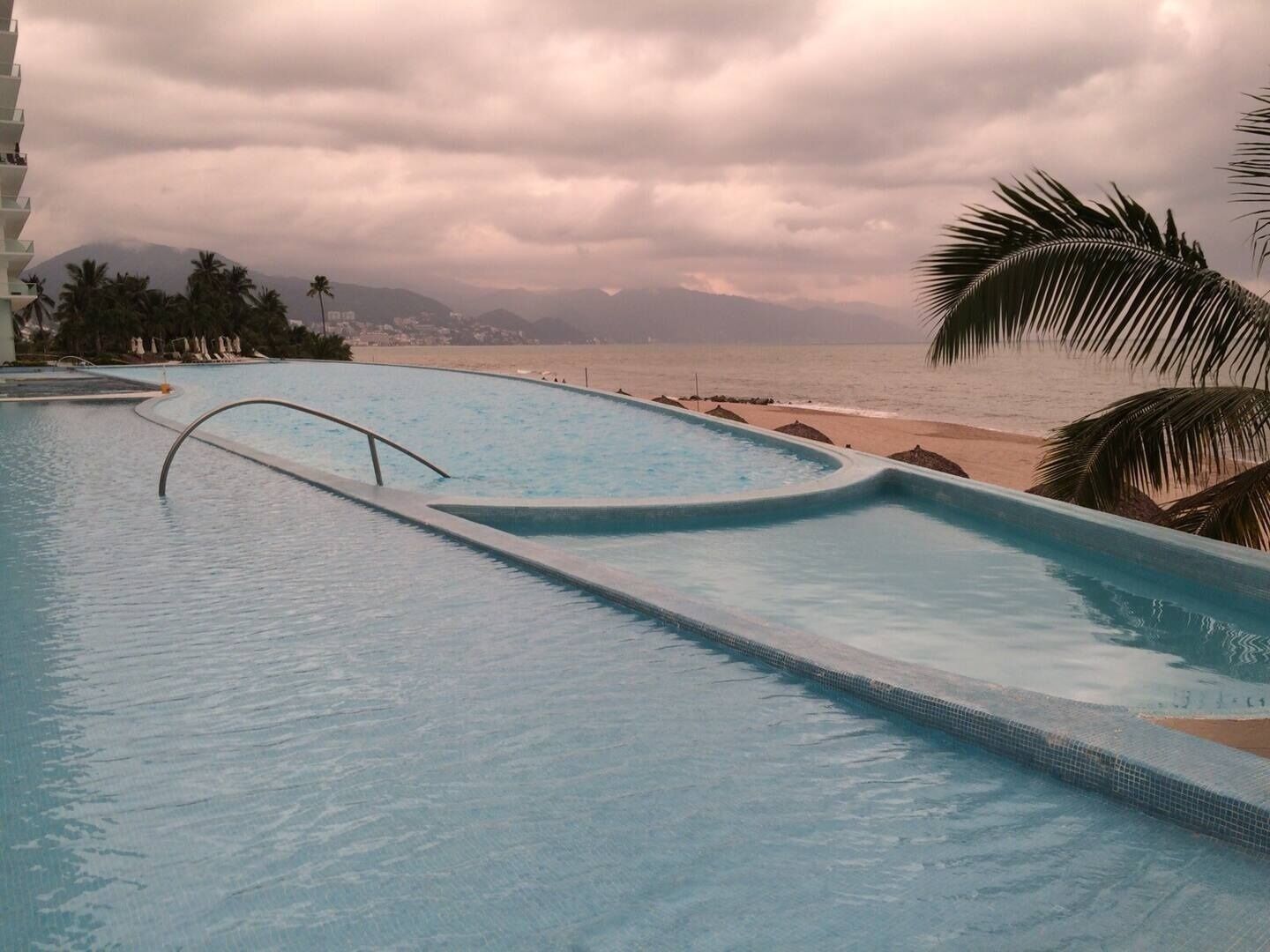 A swimming pool with a curved edge, adjacent to a beach and palm trees, under a cloudy sky.
