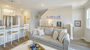Une cuisine moderne avec des meubles blancs et un réfrigérateur en acier inoxydable. On y trouve un coin repas avec des chaises blanches et une table en bois. Un canapé gris se trouve dans le salon, et une inscription sur le mur indique : « ALLEZ, PLONGEZ DANS LE LAC ! »