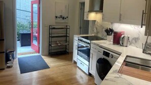 Una cocina compacta con horno empotrado, microondas y lavadora, una puerta con marco rojo y un pequeño espacio exterior visible a través de la puerta.