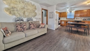 A living room with a sofa, patterned cushions, a raccoon wall decoration, and a kitchen with wooden cabinets and a dining area.